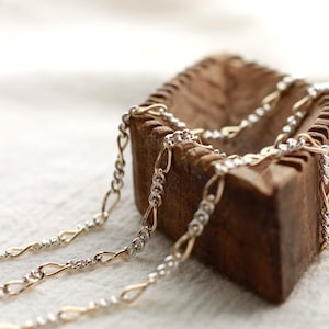 May include: Close-up of two-tone gold and silver chain necklaces. The chains are draped over a rustic, wooden container. The chains have a delicate, interwoven design. The background is a soft, white fabric.