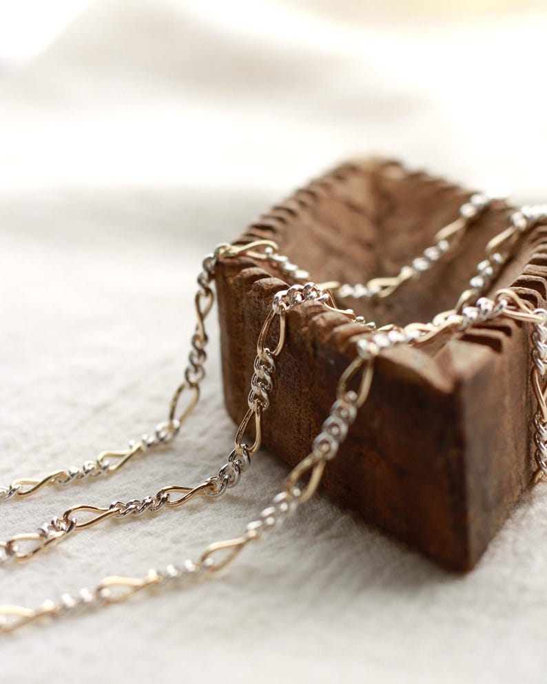 May include: Close-up of two-tone gold and silver chain necklaces. The chains are draped over a rustic, wooden container. The chains have a delicate, interwoven design. The background is a soft, white fabric.