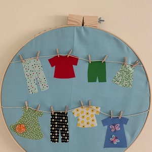 May include: A blue fabric circle with a wooden frame, featuring a clothesline with various patterned fabric clothes hanging from it. The clothes include a red shirt, green pants, a floral dress, and patterned pants.