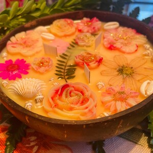 Op de afbeelding: Een gele kaars in een houten kom met een bloemmotief. De kaars heeft roze, witte en gouden bloemen, schelpen en varenbladeren.