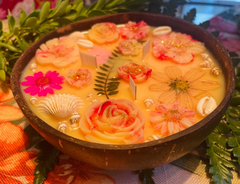 Op de afbeelding: Een gele kaars in een houten kom met een bloemmotief. De kaars heeft roze, witte en gouden bloemen, schelpen en varenbladeren.