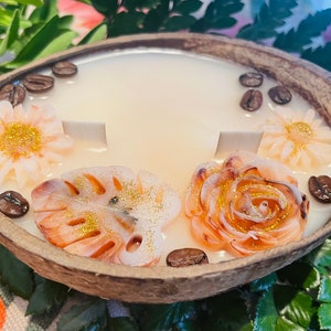 Puede incluir: Una vela de c&aacute;scara de coco con una base de cera blanca y elementos decorativos. La vela presenta una rosa, una hoja de monstera y dos margaritas, todas con detalles de purpurina dorada. Granos de caf&eacute; est&aacute;n esparcidos alrededor de la vela.