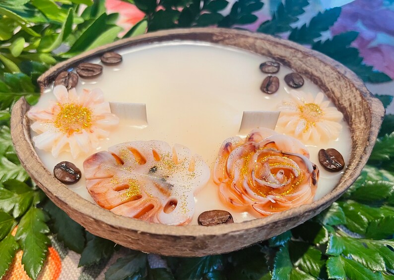 Puede incluir: Una vela de c&aacute;scara de coco con una base de cera blanca y elementos decorativos. La vela presenta una rosa, una hoja de monstera y dos margaritas, todas con detalles de purpurina dorada. Granos de caf&eacute; est&aacute;n esparcidos alrededor de la vela.