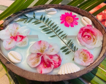 May include: A coconut shell candle with a light blue wax base and pink and white flowers embedded in the wax. The candle is decorated with seashells and a green leaf.