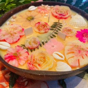 Op de afbeelding: Een ronde houten kom gevuld met een gele kaars. De kaars is versierd met roze, witte en gouden bloemen, schelpen en bladeren.