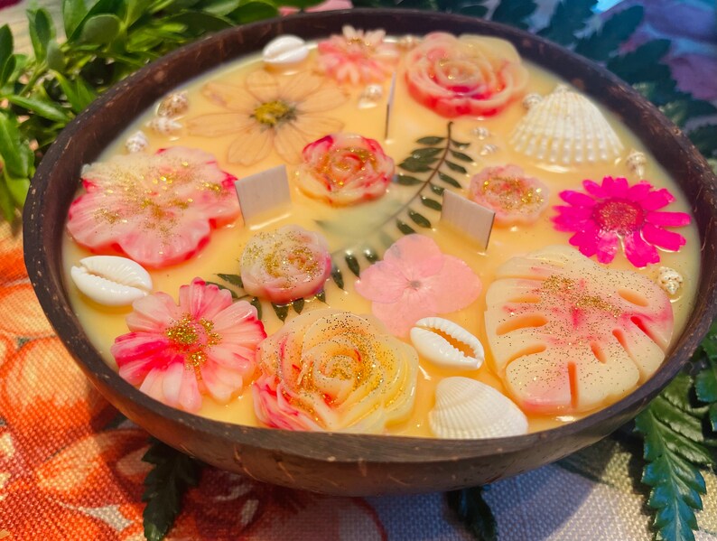 Op de afbeelding: Een ronde houten kom gevuld met een gele kaars. De kaars is versierd met roze, witte en gouden bloemen, schelpen en bladeren.
