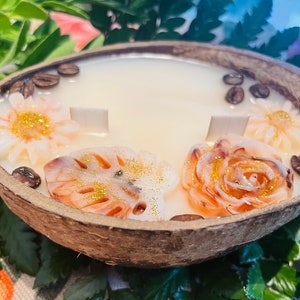 Puede incluir: Una vela de c&aacute;scara de coco con una base de cera blanca y detalles decorativos florales y de hojas. La vela est&aacute; decorada con purpurina dorada y granos de caf&eacute;.