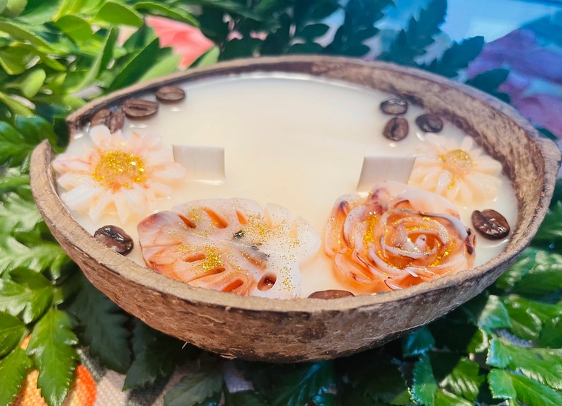 Puede incluir: Una vela de c&aacute;scara de coco con una base de cera blanca y detalles decorativos florales y de hojas. La vela est&aacute; decorada con purpurina dorada y granos de caf&eacute;.