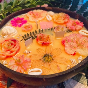 Op de afbeelding: Een ronde houten kom gevuld met een gele kaars. De kaars is versierd met roze en witte bloemen, schelpen en gouden glitter.