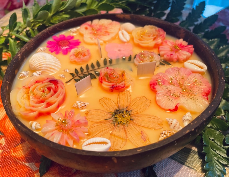 Op de afbeelding: Een ronde houten kom gevuld met een gele kaars. De kaars is versierd met roze en witte bloemen, schelpen en gouden glitter.