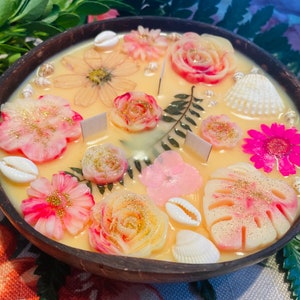 Op de afbeelding: Een kokosnootschelpkaars met een bloemen- en schelpdesign. De kaars is lichtgeel van kleur met roze, witte en gouden accenten. De kaars is versierd met schelpen, bloemen en bladeren.