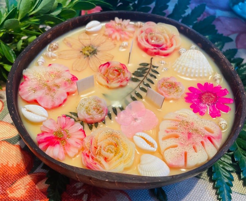 Op de afbeelding: Een kokosnootschelpkaars met een bloemen- en schelpdesign. De kaars is lichtgeel van kleur met roze, witte en gouden accenten. De kaars is versierd met schelpen, bloemen en bladeren.