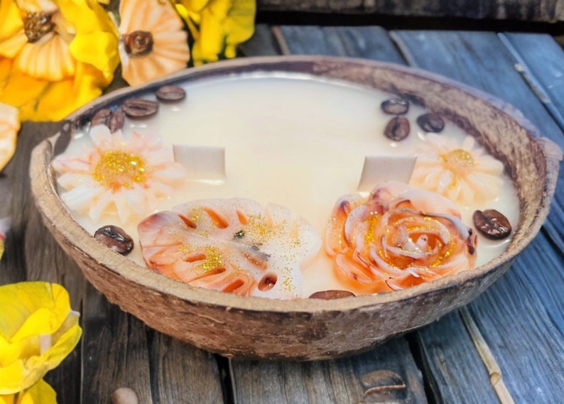 Puede incluir: Una vela de c&aacute;scara de coco con una base de cera blanca y tres elementos decorativos: una flor, una hoja y una rosa. La vela est&aacute; decorada con granos de caf&eacute; y purpurina dorada.