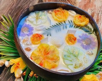 May include: A coconut shell candle with a white base and various floral and leaf designs. The candle features a purple flower, a yellow flower, a green leaf, and a white seashell.