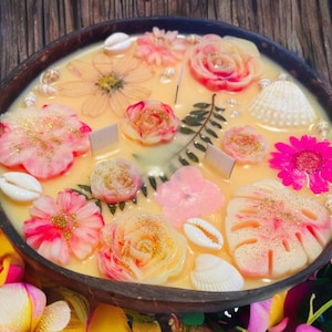 Op de afbeelding: Een ronde, gele kaars met verschillende geperste bloemen en schelpen. De kaars bevindt zich in een houten kom en heeft een roze, wit en gouden kleurenschema.