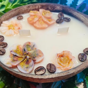 Puede incluir: Una vela de soja blanca en una c&aacute;scara de coco con tres mechas. La vela est&aacute; decorada con tres flores rosas y blancas y granos de caf&eacute;.