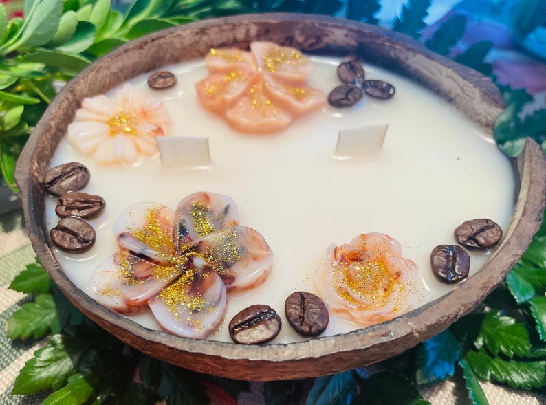 Puede incluir: Una vela de soja blanca en una c&aacute;scara de coco con tres mechas. La vela est&aacute; decorada con tres flores rosas y blancas y granos de caf&eacute;.