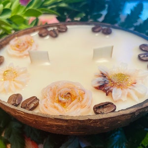 Puede incluir: Una vela blanca con un dise&ntilde;o floral y granos de caf&eacute; alrededor del borde, colocada en una c&aacute;scara de coco natural.