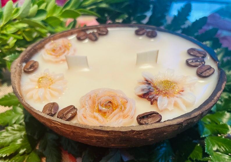 Puede incluir: Una vela blanca con un dise&ntilde;o floral y granos de caf&eacute; alrededor del borde, colocada en una c&aacute;scara de coco natural.
