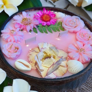 May include: A pink soy candle in a coconut shell container with a floral design. The candle features pink and white flowers, seashells, and gold glitter.