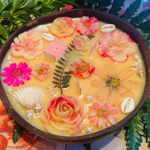 Op de afbeelding: Een ronde houten kom gevuld met een gele kaars met verschillende geperste bloemen en schelpen. De kaars heeft een bloemmotief met roze, witte en gele bloemen.