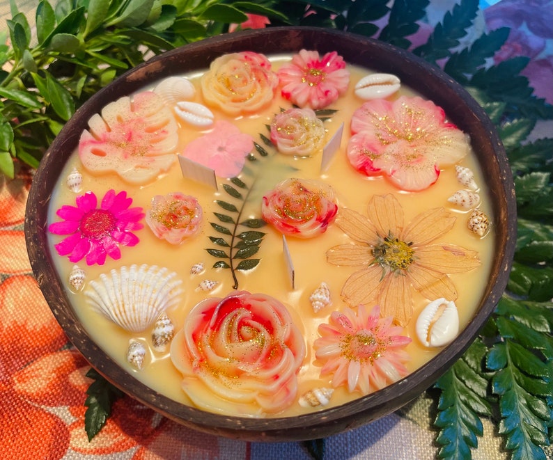 Op de afbeelding: Een ronde houten kom gevuld met een gele kaars met verschillende geperste bloemen en schelpen. De kaars heeft een bloemmotief met roze, witte en gele bloemen.