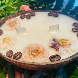 Puede incluir: Una vela blanca con un dise&ntilde;o floral y granos de caf&eacute; en una c&aacute;scara de coco. La vela tiene tres mechas y est&aacute; decorada con tres flores y granos de caf&eacute;.
