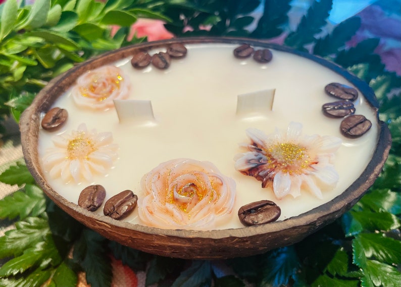 Puede incluir: Una vela blanca con un dise&ntilde;o floral y granos de caf&eacute; en una c&aacute;scara de coco. La vela tiene tres mechas y est&aacute; decorada con tres flores y granos de caf&eacute;.