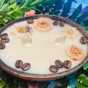 Puede incluir: Una vela blanca con un dise&ntilde;o floral y de granos de caf&eacute; en una c&aacute;scara de coco natural. La vela tiene tres mechas blancas y est&aacute; decorada con tres flores rosas y blancas y granos de caf&eacute;.