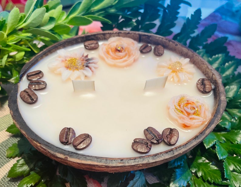 Puede incluir: Una vela blanca con un dise&ntilde;o floral y de granos de caf&eacute; en una c&aacute;scara de coco natural. La vela tiene tres mechas blancas y est&aacute; decorada con tres flores rosas y blancas y granos de caf&eacute;.