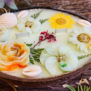 May include: A white coconut shell candle with embedded flowers and seashells. The candle features a yellow sunflower, a white daisy, a white rose, a white plumeria, and a red flower bud. The candle is decorated with gold glitter and seashells.