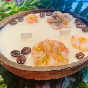 Puede incluir: Una vela de c&aacute;scara de coco con una base de cera blanca y tres decoraciones de cera en forma de flor rosa. La vela est&aacute; decorada con granos de caf&eacute; alrededor del borde.
