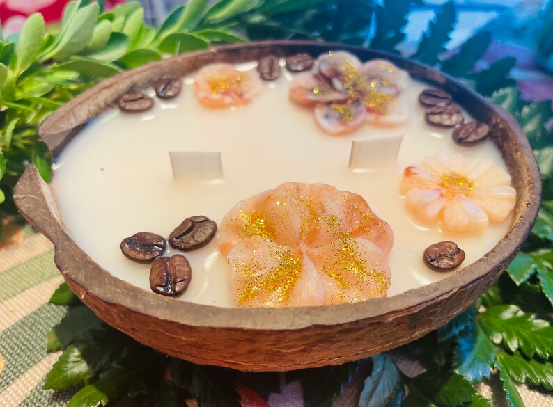 Puede incluir: Una vela de c&aacute;scara de coco con una base de cera blanca y tres decoraciones de cera en forma de flor rosa. La vela est&aacute; decorada con granos de caf&eacute; alrededor del borde.
