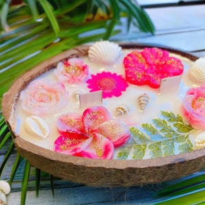 Peut inclure: Une bougie en forme de coque de noix de coco blanche avec des fleurs roses et blanches, des coquillages et de la verdure incrustés dans la cire.