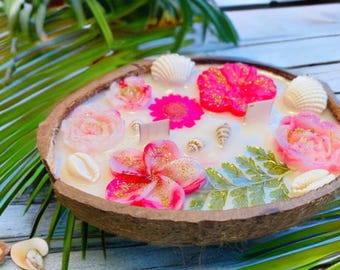May include: A white coconut shell candle with pink and white flowers, seashells, and greenery embedded in the wax.