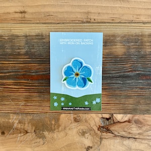 Könnte beinhalten: Ein blauer gestickter Vergissmeinnicht-Blumen-Patch mit einem Bügeleisen-Rücken. Der Patch befindet sich auf einer weißen Karte mit dem Text "Embroidered Patch with Iron-On Backing" und der Website "wrenandtheraven.com".