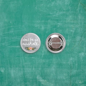 Op de afbeelding: Twee ronde, grijze knopen op een groene ondergrond. Eén knoop heeft de tekst "don't be an asshole" in witte letters, met een bloemenontwerp. De andere knoop toont de achterkant van de speld. De knopen zijn van metaal.