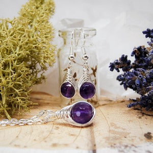 Dark purple amethyst bead jewellery wire wrapped in a herringbone design in silver wire, dangle earrings hang from a glass vial with a matching pendant laying in front.