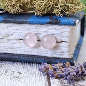 Peut inclure: Deux boucles d'oreilles rondes en quartz rose pâle. Les boucles d'oreilles sont présentées sur une surface en bois blanc et gris, à côté d'un livre ancien avec un dos bleu et des brins de lavande séchée.