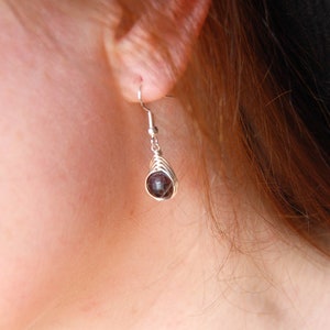 A dark plum red faceted garnet bead wrapped in a herringbone design in silver wire creates a beautiful dangle earring that is hanging from the models ear lobe.