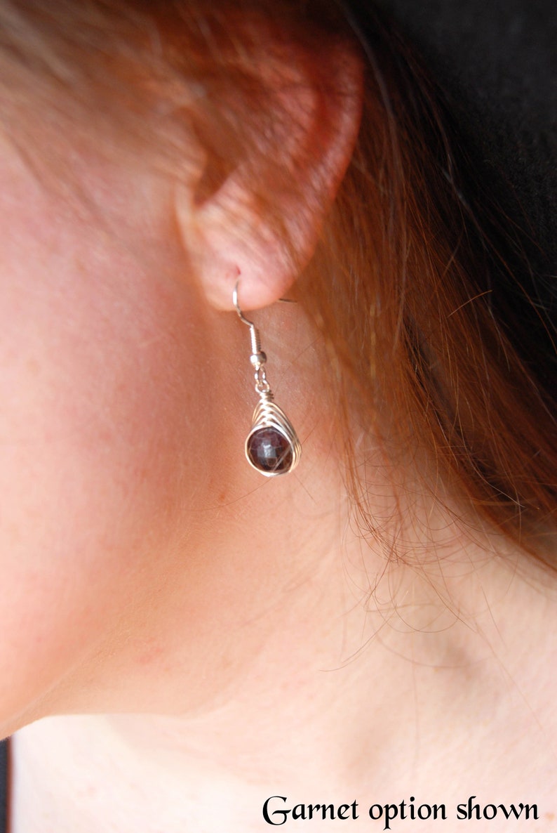 A dark plum red faceted garnet bead wrapped in a herringbone design in silver wire creates a beautiful dangle earring that is hanging from the models ear lobe.