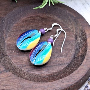 May include: A pair of shell earrings. Each earring has a vibrant, multicolored shell design with blue, yellow, and purple hues. The shells hang from silver-coloured hooks and are accented with a small purple bead. The earrings are displayed on a wooden surface.