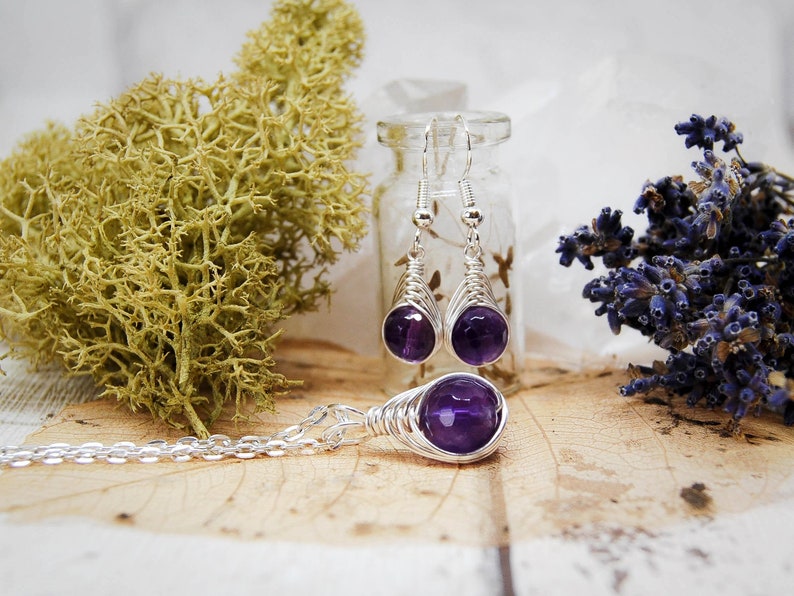 Dark purple amethyst bead jewellery wire wrapped in a herringbone design in silver wire, dangle earrings hang from a glass vial with a matching pendant laying in front.