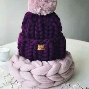 May include: A purple knitted beanie with a fluffy pink pom pom and a matching pink knitted band. The beanie has a brown label that says "Your Yarn".