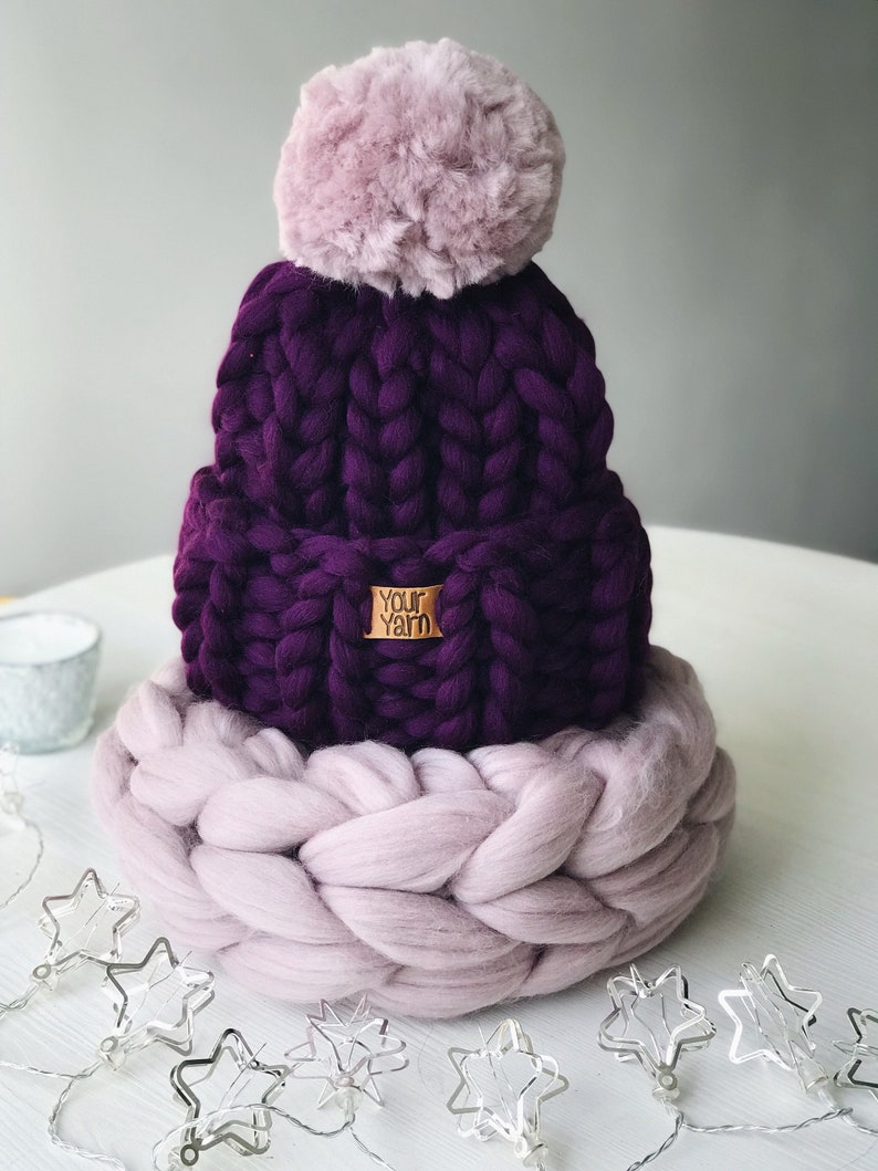 May include: A purple knitted beanie with a fluffy pink pom pom and a matching pink knitted band. The beanie has a brown label that says "Your Yarn".