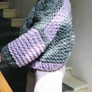 May include: A chunky knit sweater with a striped pattern in shades of grey and lavender. The sweater has a relaxed fit and a cropped length.