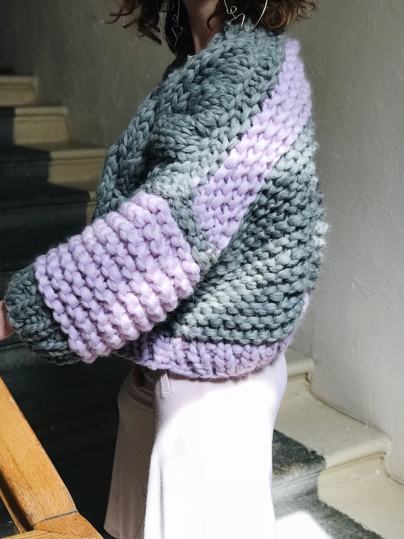 May include: A chunky knit sweater with a striped pattern in shades of grey and lavender. The sweater has a relaxed fit and a cropped length.