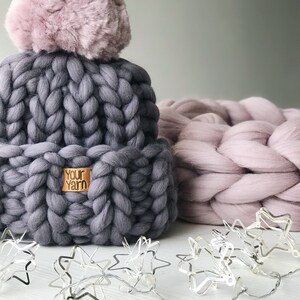 May include: A gray chunky knit beanie with a pink pom pom and a brown leather tag that says "Your Yarn". The beanie is sitting on a white surface with a pink chunky knit blanket and a string of silver star lights.
