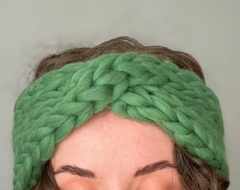 May include: A green knitted headband with a twisted design. The headband is made of thick yarn and has a soft, cozy feel.