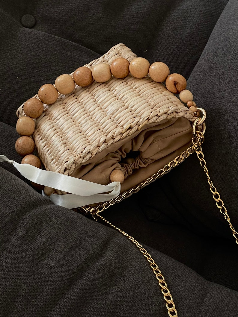 May include: A small straw bag with a wooden bead handle and a gold chain strap. The bag has a drawstring closure and a brown lining.